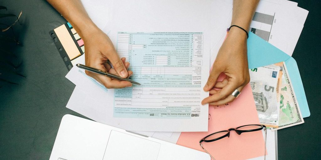 Hands writing on tax documents with laptop, glasses, and currency on desk.